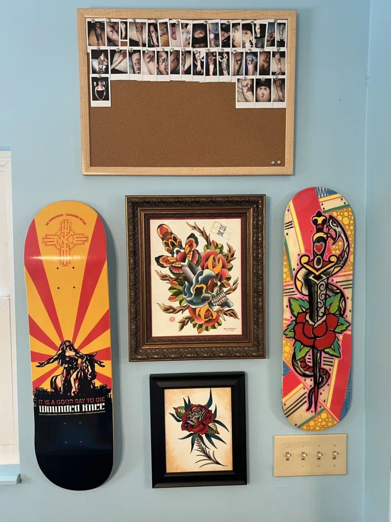 Polaroid tattoo photos pinned on a cork board, two skateboard decks, and framed tattoo art on a wall